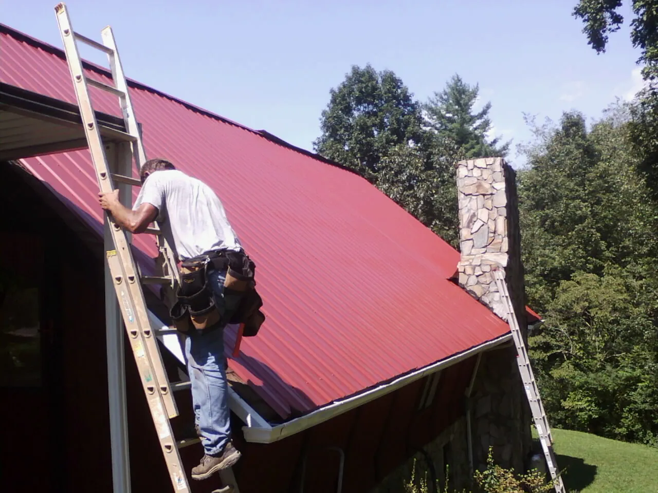 Roofer on a ladder beside a red metal roof and stone chimney — Soffit & Fascia Repair by Roofing Brothers in Absecon